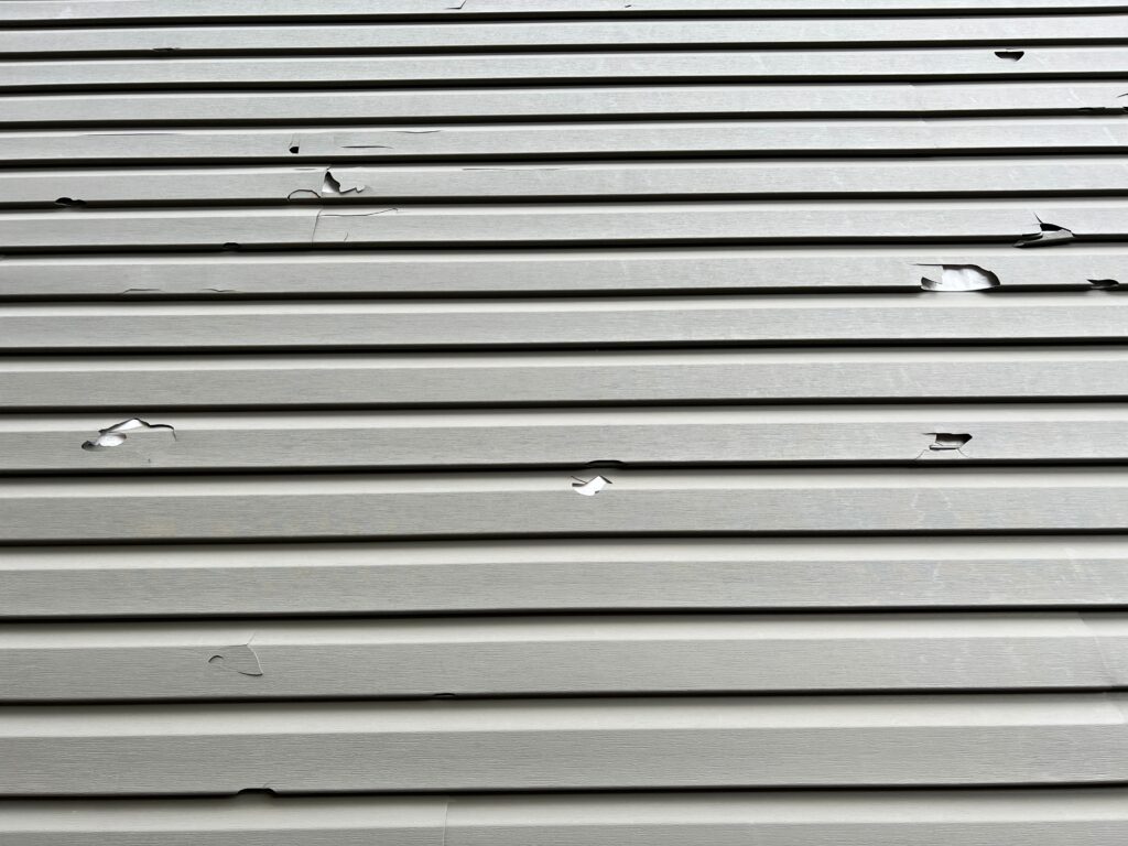 Holes in the exterior siding of the home from hail storm damage. The broken vinyl siding on a house due to hail.
