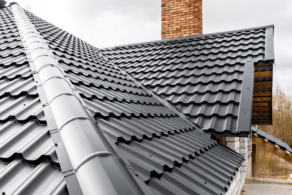 Close-up view of a dark gray metal roof with corrugated sheets and a matching rain gutter system, alongside a brick chimney, showcasing modern roofing materials and design.