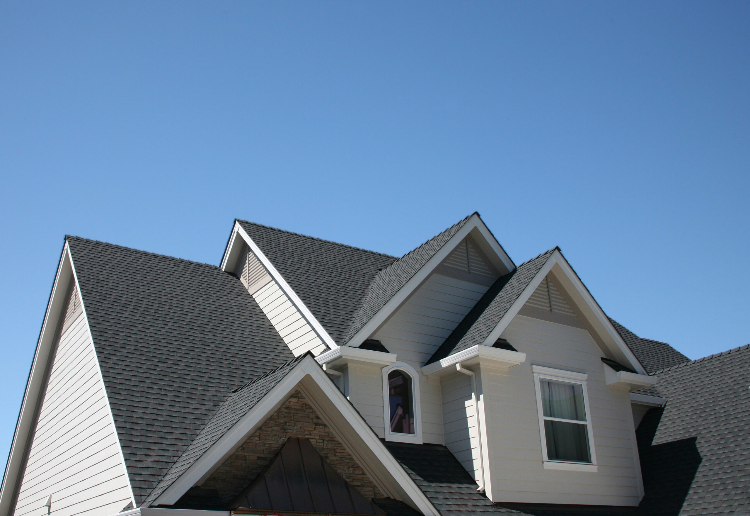 A clear blue sky provides the backdrop for a house showcasing different types of roof styles, including steep gable roofs and a dormer window, all covered in grey shingles.