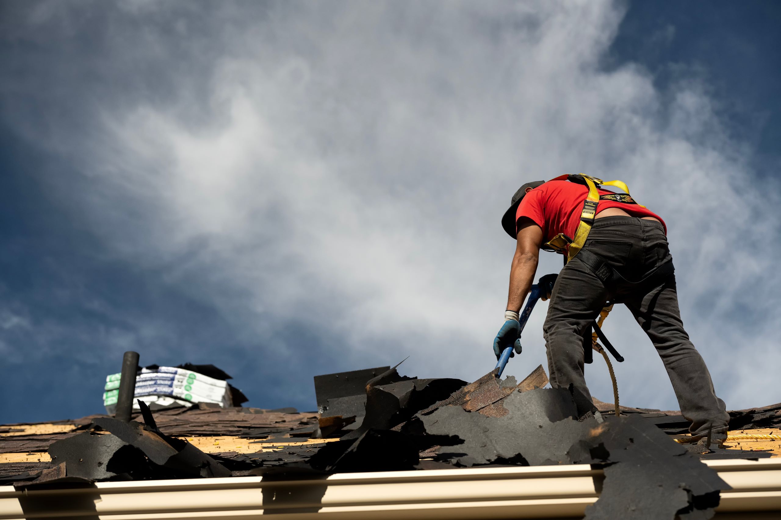 A hail damage roofing contractor in a red shirt and safety gear repairs a roof under a clear sky, removing damaged shingles caused by a recent hailstorm.